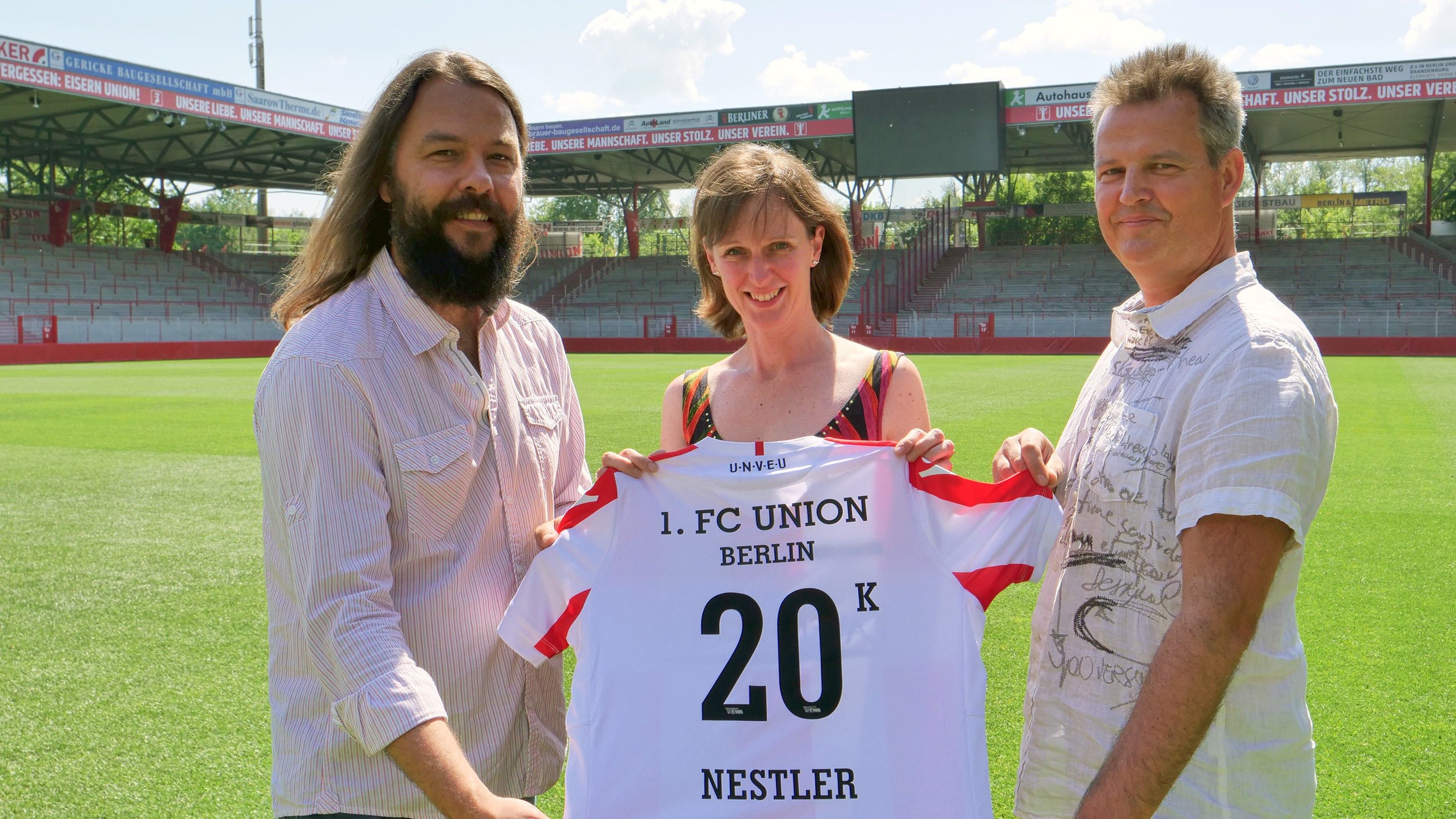 Drei Personen halten ein Fußballtrikot des 1. FC Union Berlin mit der Aufschrift "20 K Nestler" auf einem Stadionrasen.
