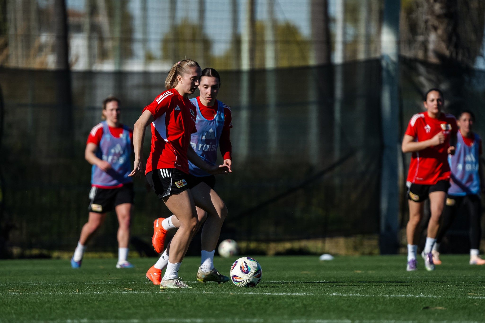 Zwei weibliche Fußballspielerinnen in roter Kleidung dribbeln den Ball auf einem Trainingsplatz, während andere Spielerinnen im Hintergrund üben.