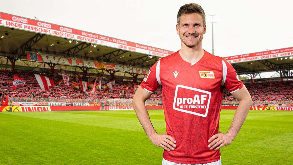 Ein Fußballspieler in rotem Trikot steht lächelnd auf dem Spielfeld eines Stadions mit zahlreichen Zuschauern im Hintergrund.