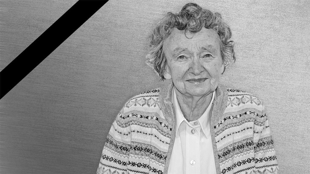 Eine ältere Frau mit lockigem Haar, lächelnd, trägt einen gestrickten Cardigan und steht vor einem grauen Hintergrund.