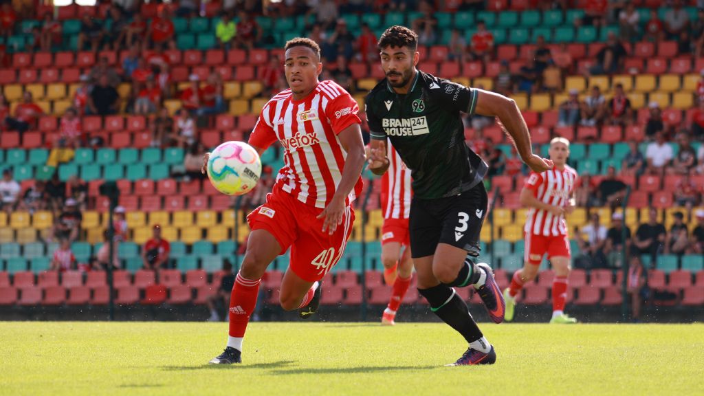 Ein Fußballspieler in roter und weißer Kleidung sprintet dem Ball hinterher, während ein Gegenspieler in dunkler Kleidung ihn verfolgt.