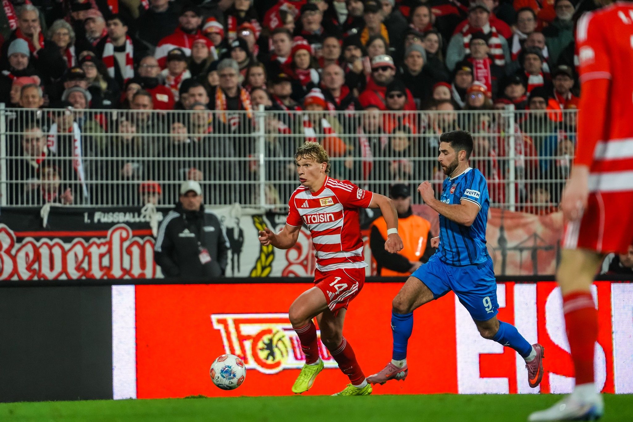 Ein Fußballspieler in rotem Trikot dribbelt den Ball, während ein gegnerischer Spieler in blauem Trikot versucht, ihn zu stoppen.