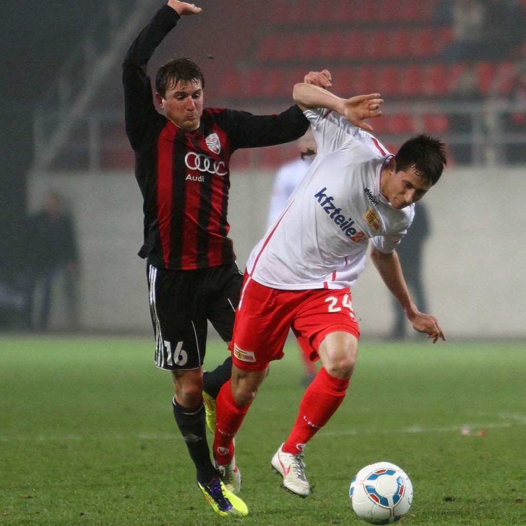 Zwei Fußballspieler im Wettkampf um den Ball; einer wird von hinten gehalten, während der andere sich freimachen möchte.