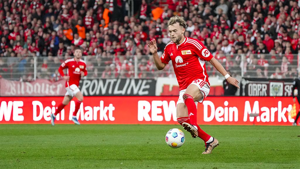 Spieler in rotem Trikot führt einen Ball auf dem Fußballfeld während eines Spiels mit jubelnden Fans im Hintergrund.