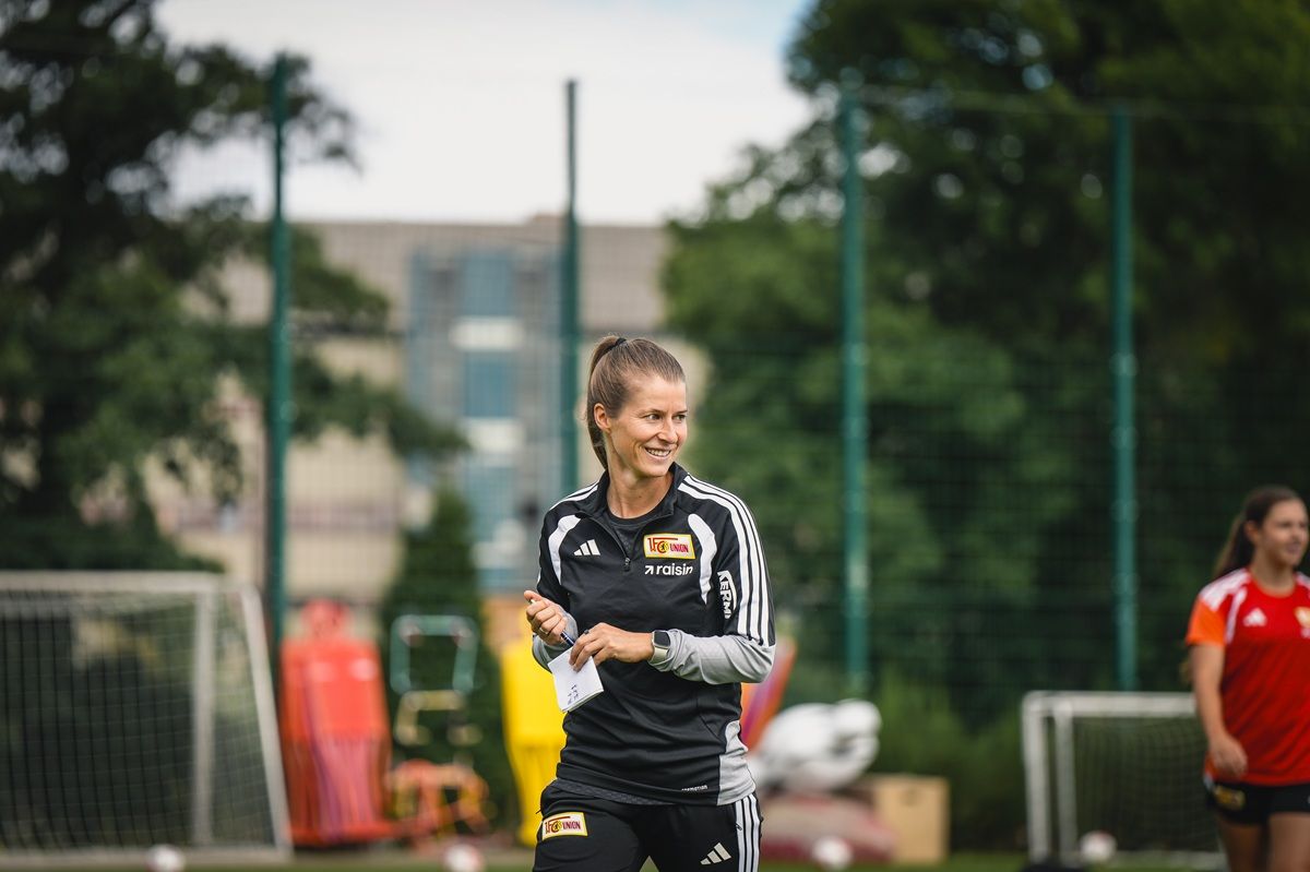 Eine Trainerin steht auf einem Sportplatz, lächelt und hält Notizen in der Hand, im Hintergrund sind Sportgeräte sichtbar.