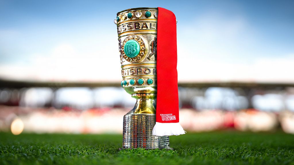 A magnificent football trophy with golden and turquoise accents stands on green grass, with a red scarf beside it.