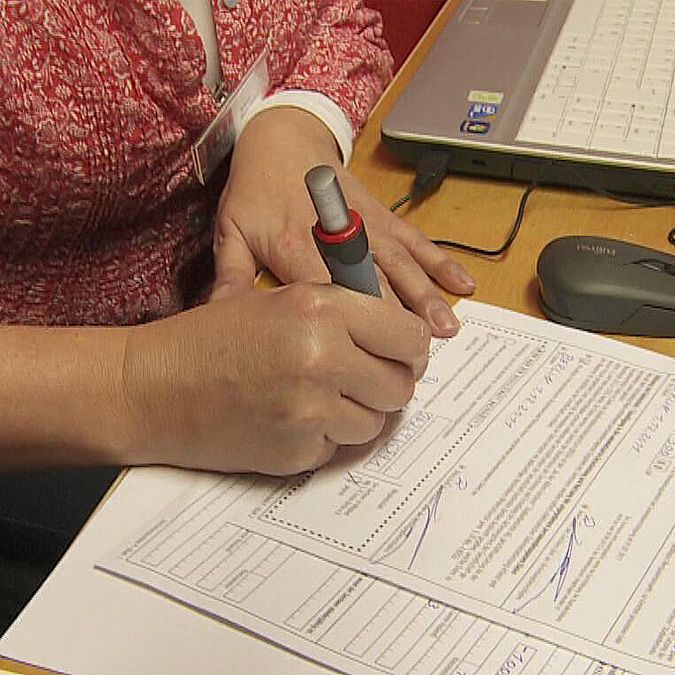 Eine Hand hält einen Stift und signiert ein Dokument, während ein Laptop und eine Maus im Hintergrund zu sehen sind.