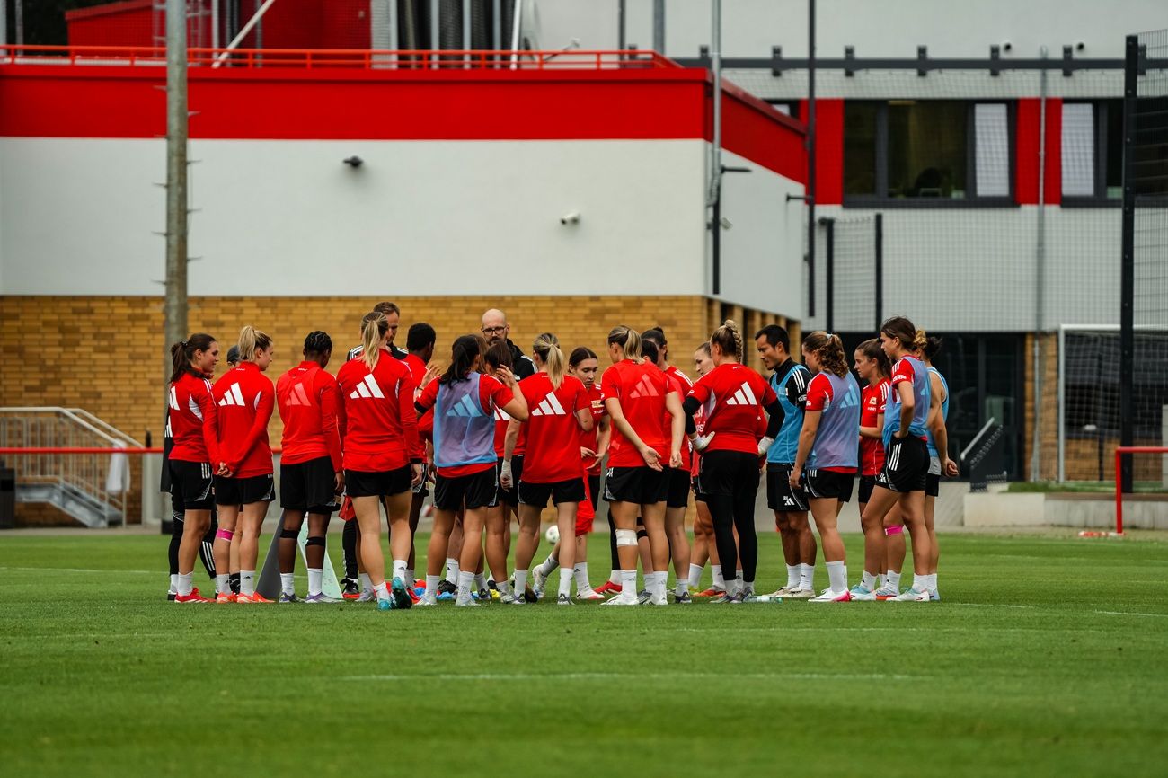 Gruppentraining einer Fußballmannschaft mit Spielerinnen in roten Trikots, die sich auf einem Trainingsplatz versammeln.