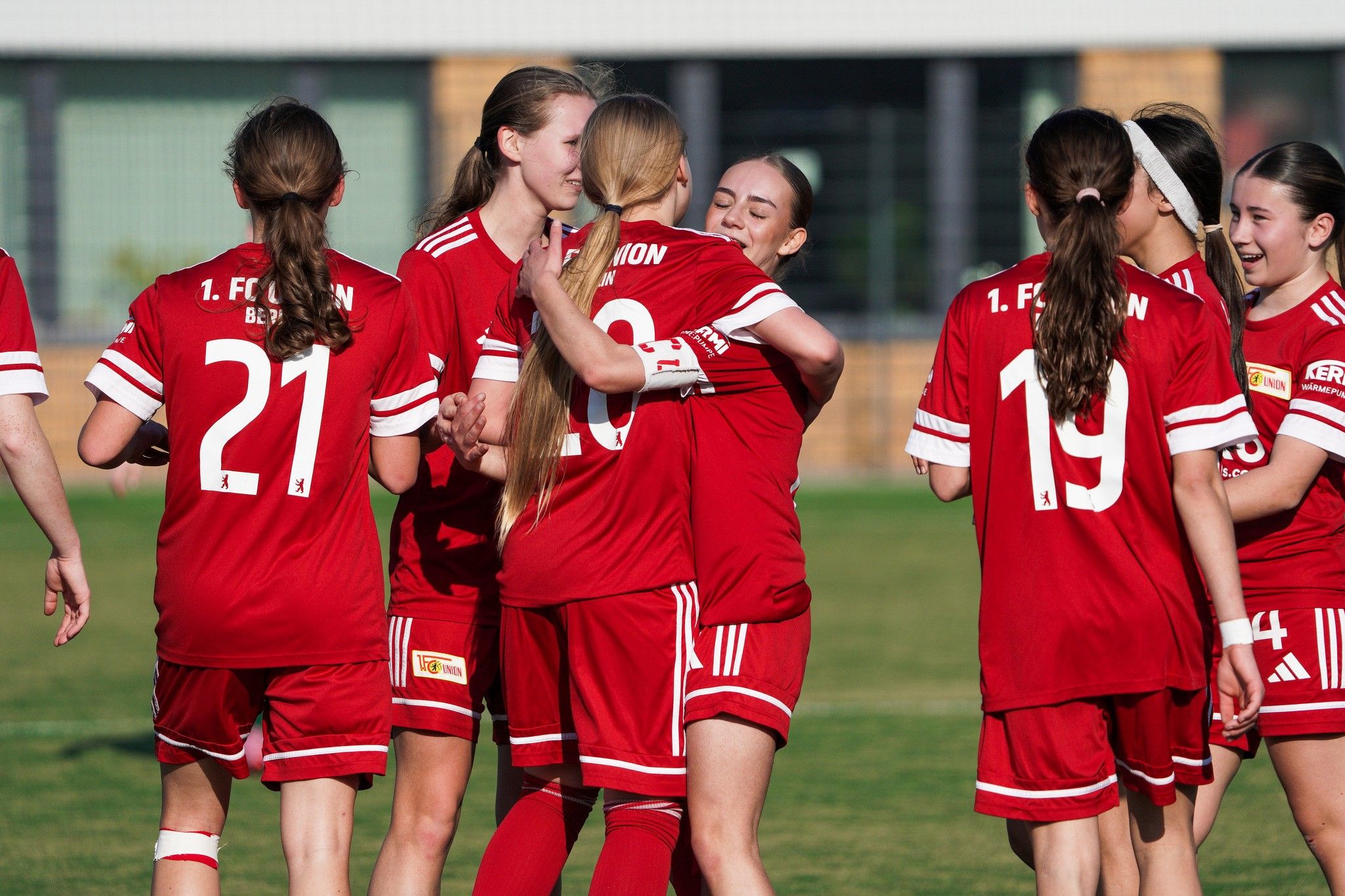 Mannschaft in roten Fußballtrikots jubelt und umarmt sich auf dem Spielfeld, Sonnenlicht scheint auf die Szene.