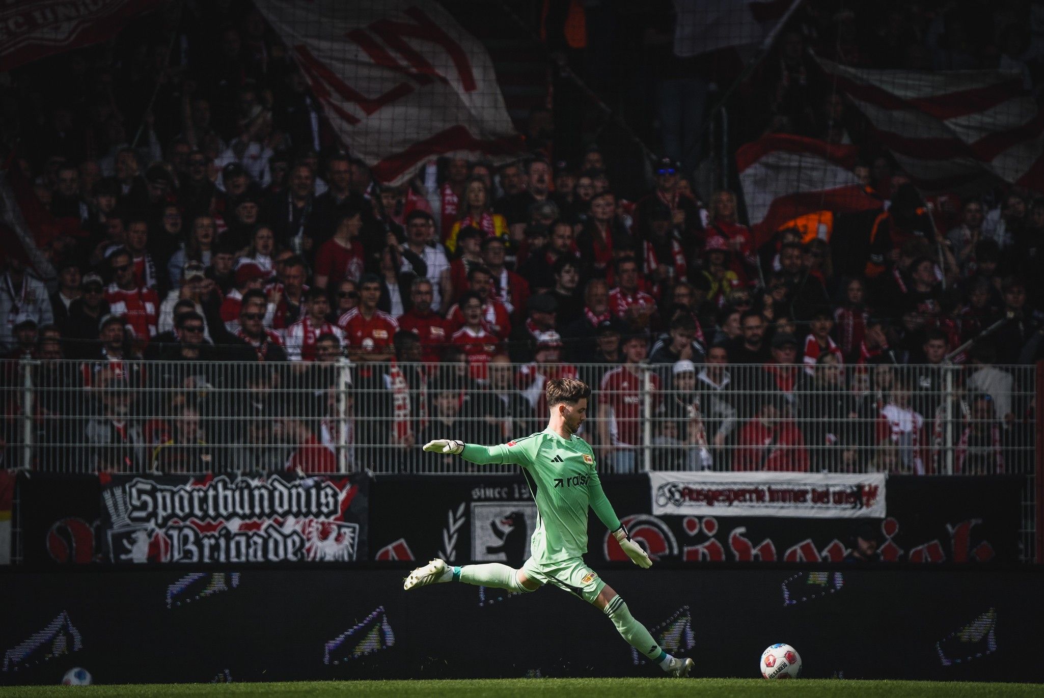 Torwart springt in die Luft, während er einen Ball im Stadion abwehrt. Im Hintergrund Banner und jubelnde Fans.