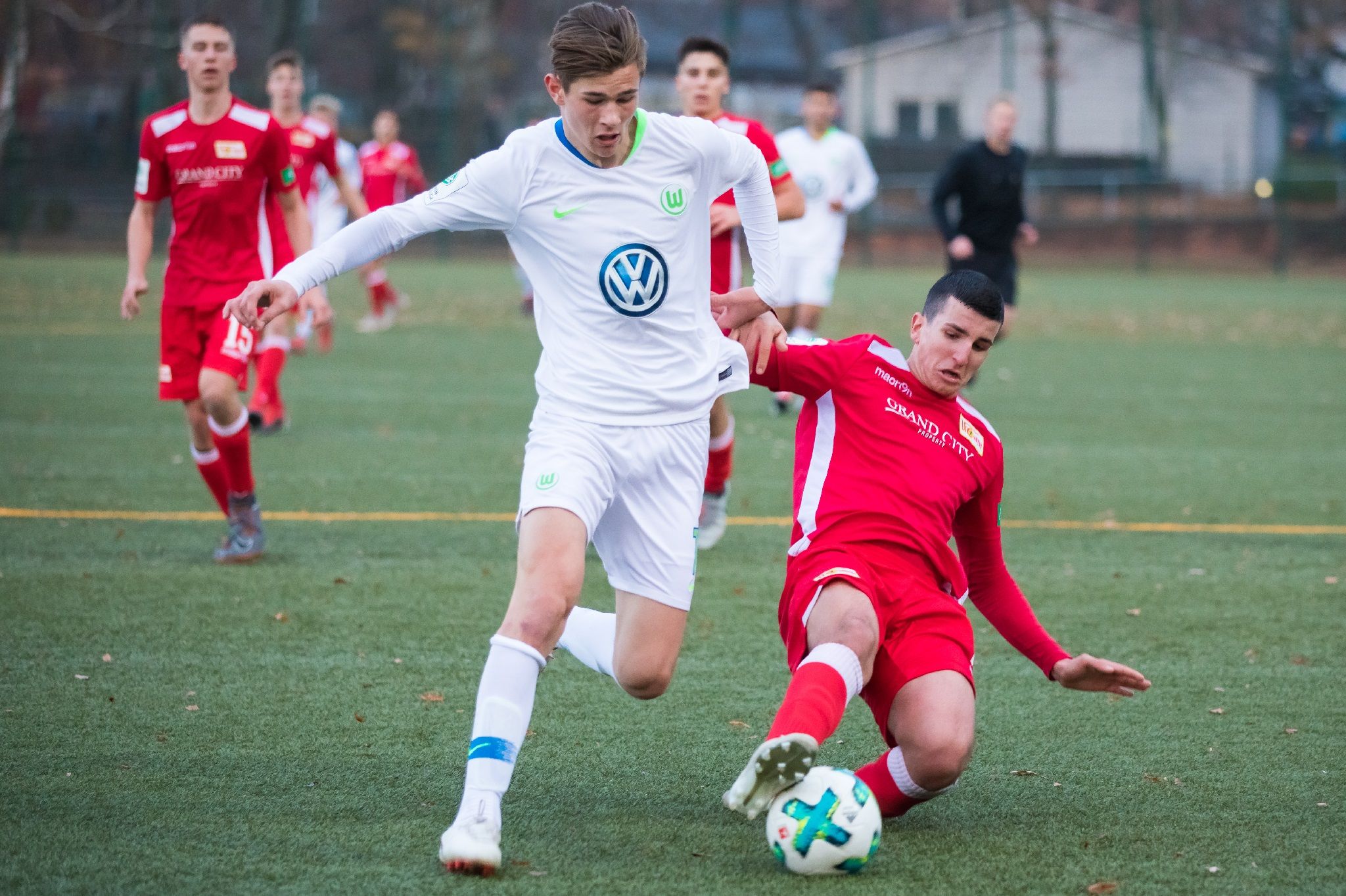 Ein Fußballspieler in weißem Trikot dribbelt den Ball, während ein Gegner in rotem Trikot ihn zu stoppen versucht.