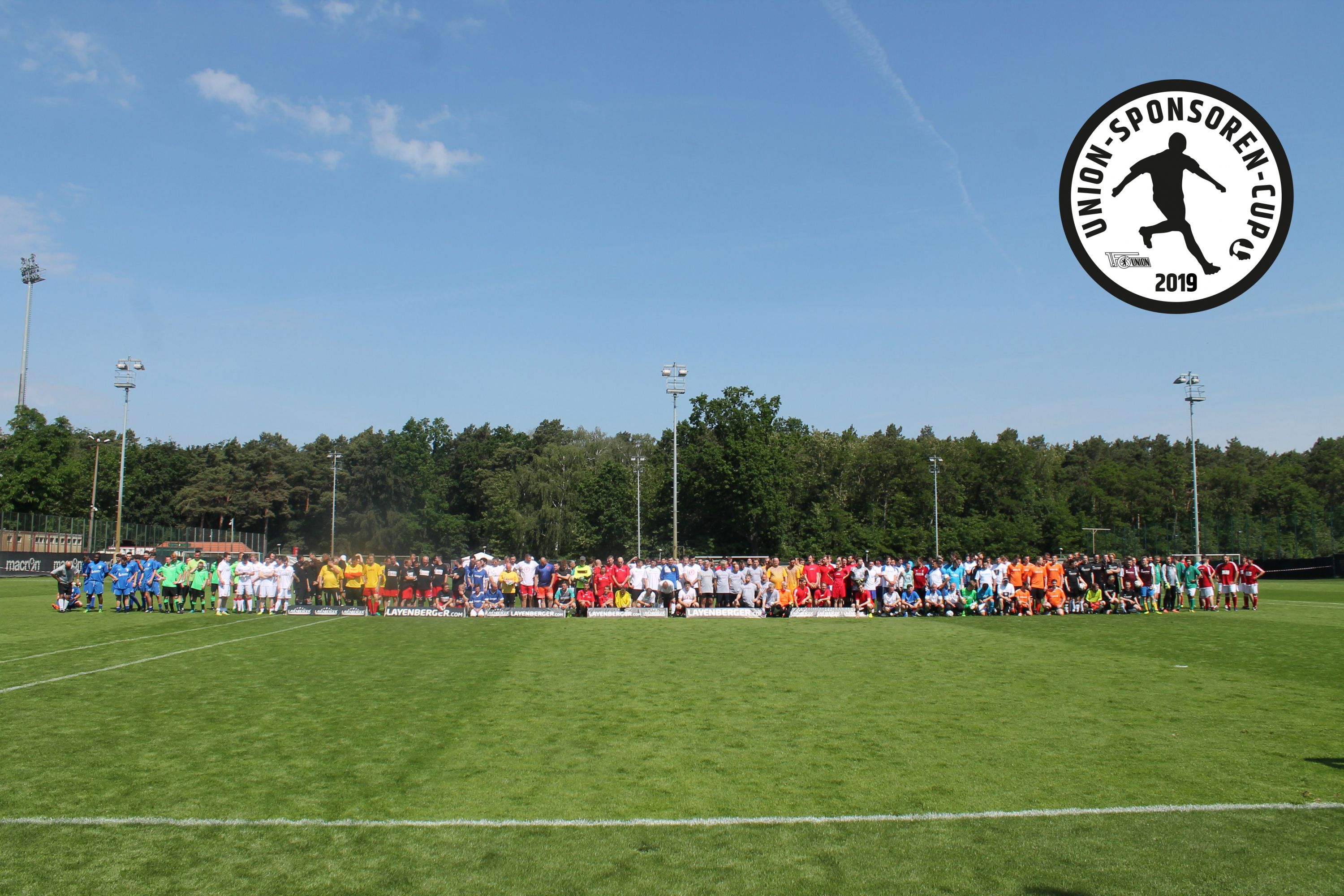 Gruppierung von Spielern in verschiedenen Trikots auf einem Fußballfeld, umgeben von Bäumen und unter blauem Himmel.