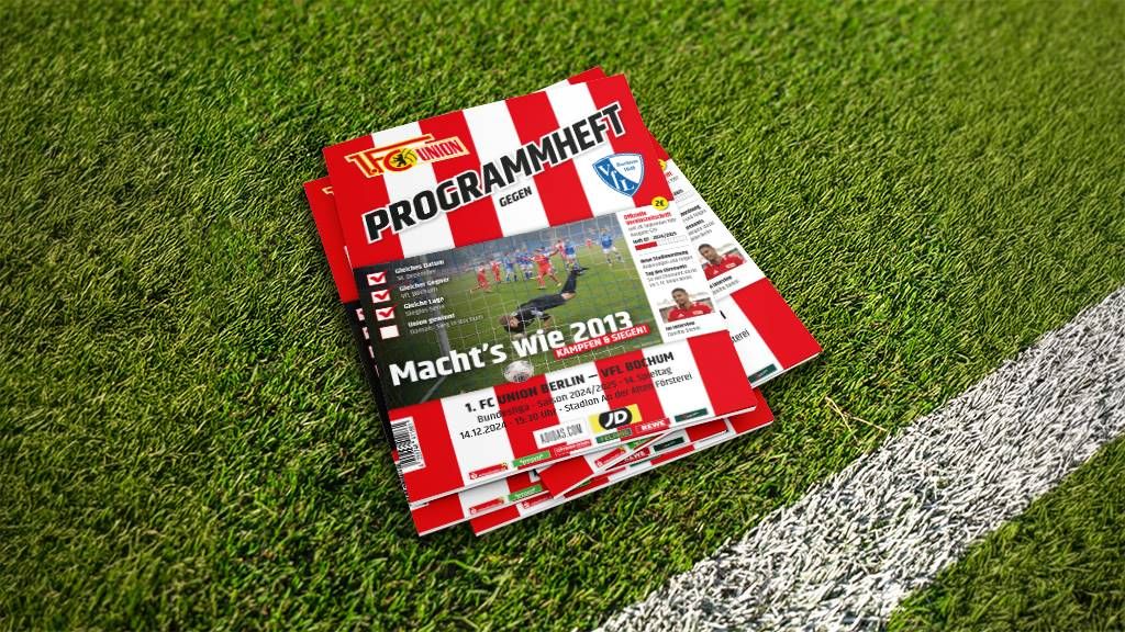 Ein Stapel Programmhefte mit Vereinsfarben auf einem grünen Rasen, Teil eines Fußballfeldes.