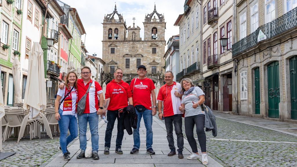 Six people in red T-shirts are standing on a paved street in a city, with historical buildings in the background.