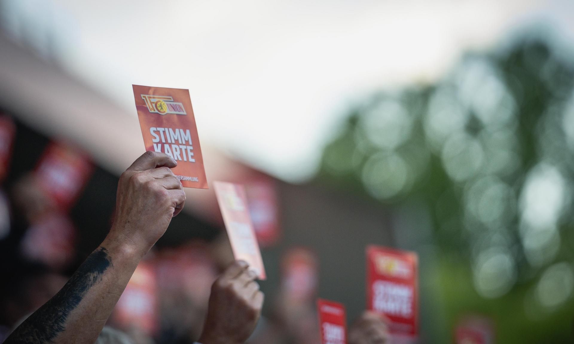 Mehrere Hände halten rot-orangefarbene Stimmkarten in die Luft. Im Hintergrund sind verschwommene Personen und Bäume zu sehen.
