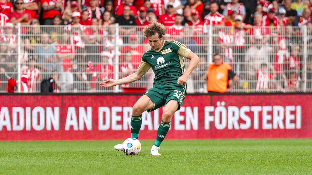 Ein Fußballspieler in grünem Trikot schießt den Ball, während ein begeistertes Publikum im Hintergrund zuschaut.