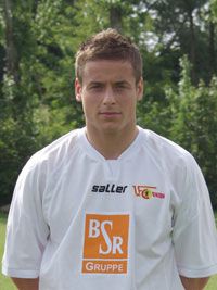 Ein junger Mann in einem weißen Fußballtrikot mit Logo steht im Freien vor grüner Umgebung.