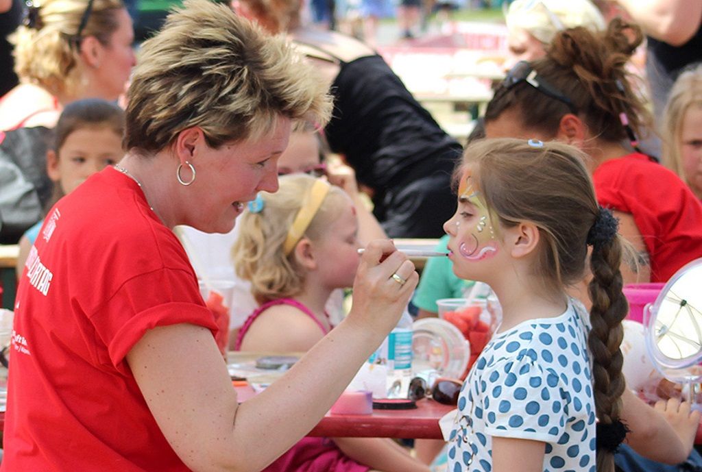 Eine Frau schminkt ein Mädchen mit bunten Farben auf einem Kinderfest. Hintergrund zeigt weitere Kinder und Aktivitäten.