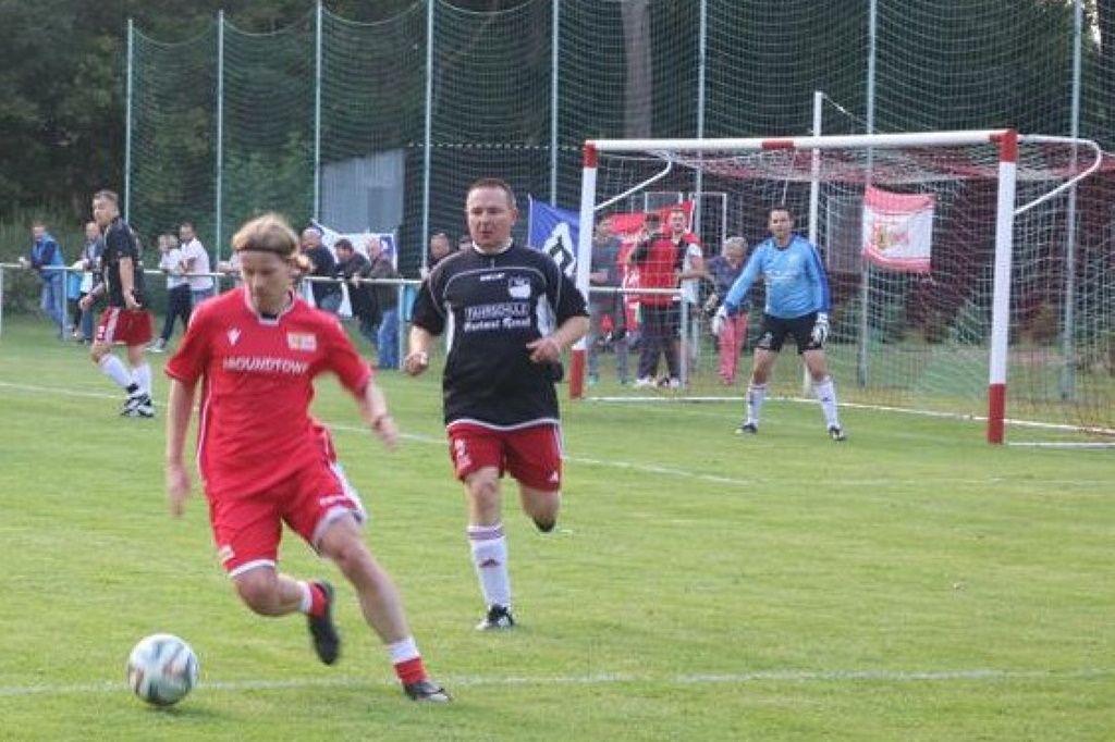 Ein Fußballspiel auf einem Rasenplatz mit Spielern in roten und schwarzen Trikots, einem Tor im Hintergrund und Zuschauern.
