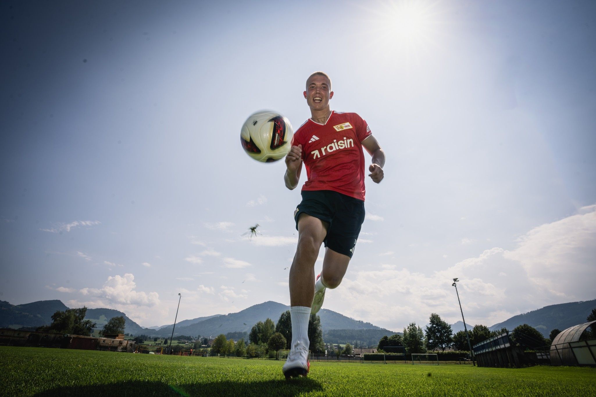 Ein junger Fußballspieler in rotem Trikot läuft mit einem Ball auf einem grünen Platz, im Hintergrund sind Berge und ein blauer Himmel.