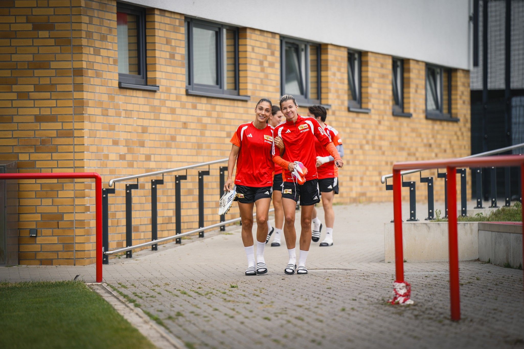 Zwei Fußballspielerinnen lächeln und gehen auf einem Weg zu einem Gebäude, während weitere Spielerinnen im Hintergrund folgen.