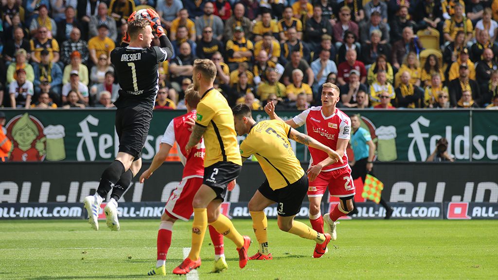 Ein Torwart in schwarzer Kleidung fängt den Ball, während Spieler in gelben und roten Trikots im Hintergrund stehen. Zuschauer sind im Stadion zu sehen.