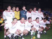 Eine Fußballmannschaft posiert jubelnd in einheitlichen weißen Trikots mit roten Akzenten, umgeben von Cheerleadern.