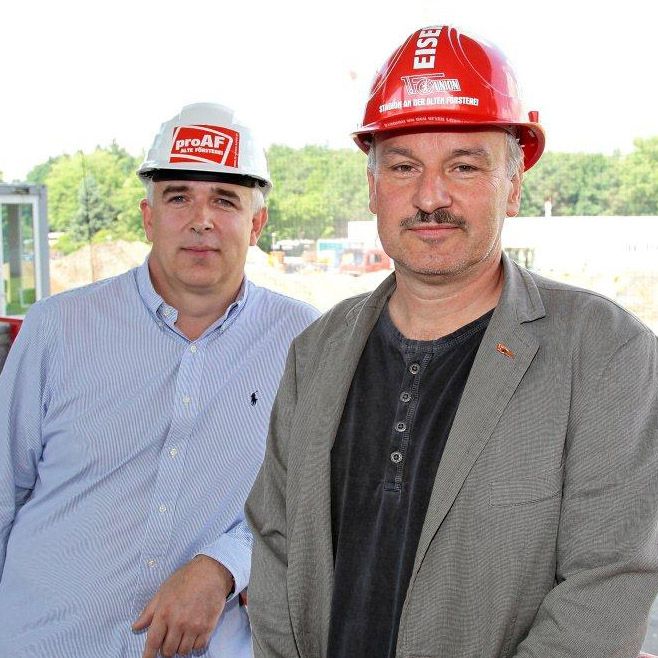 Zwei Männer stehen auf einer Baustelle, einer trägt einen weißen Helm mit Logo, der andere einen roten Sicherheitshelm.