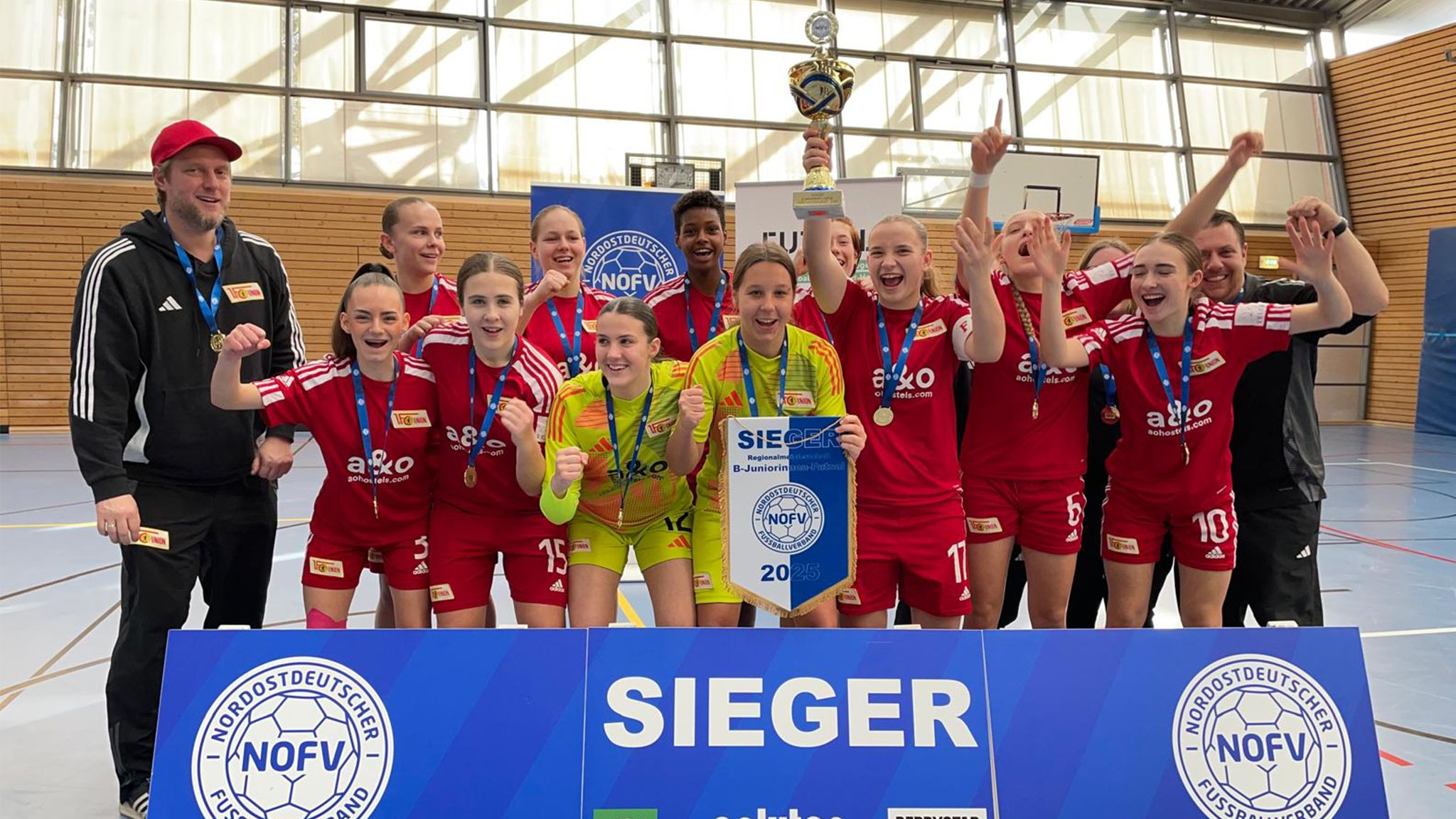 Die jubelnde Frauenmannschaft in roten Trikots hält einen Pokal und posiert vor einer Siegertafel in einer Sporthalle.