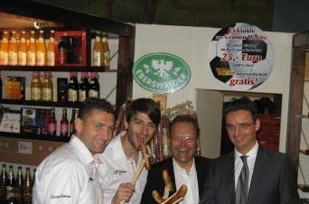 Vier Männer stehen in einem Raum mit Getränken im Hintergrund, während sie Würstchen halten und lächeln.