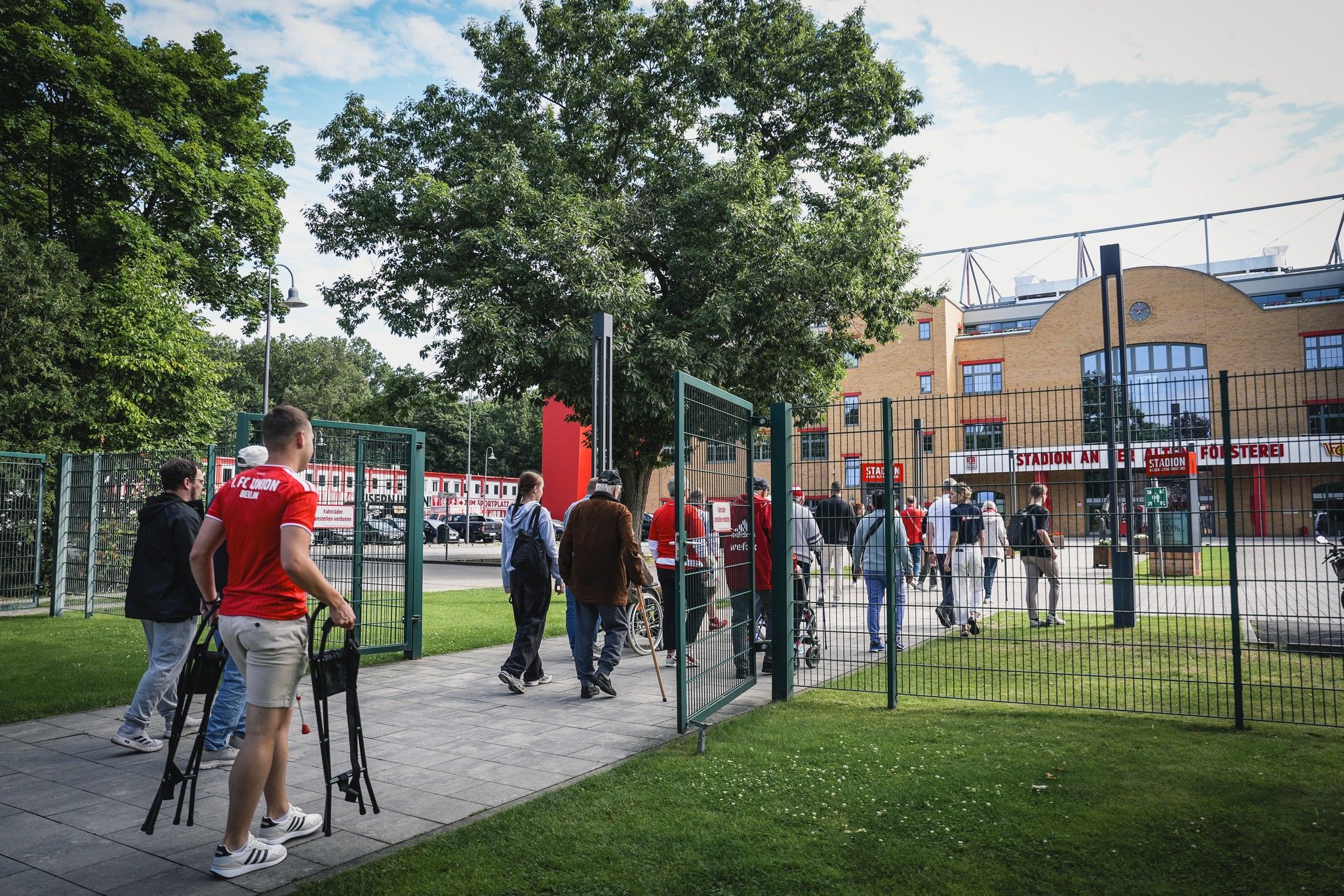 Gruppe von Menschen, die auf einen Eingang eines Stadions zugeht, umgeben von Bäumen und Wiese.