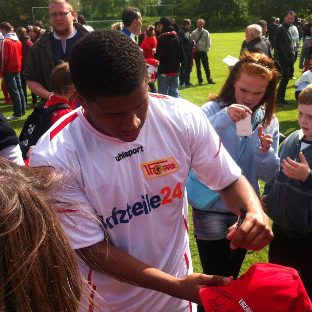 Ein Fußballspieler signiert einen roten Hut, umgeben von Fans in einer Sportumgebung.
