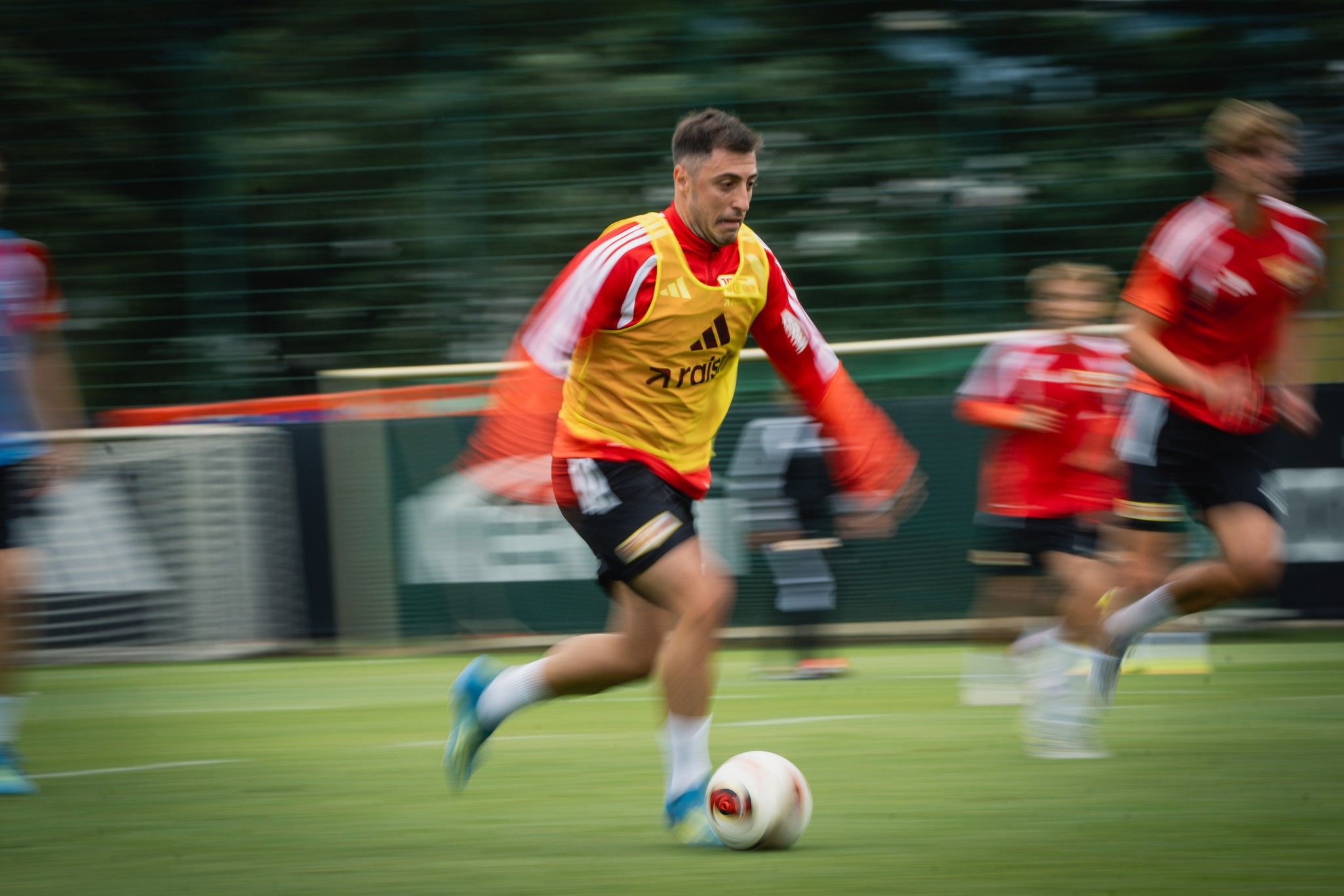 Ein Fußballspieler in Gelb bei einem Training. Er läuft schnell, während andere Spieler im Hintergrund zu sehen sind.