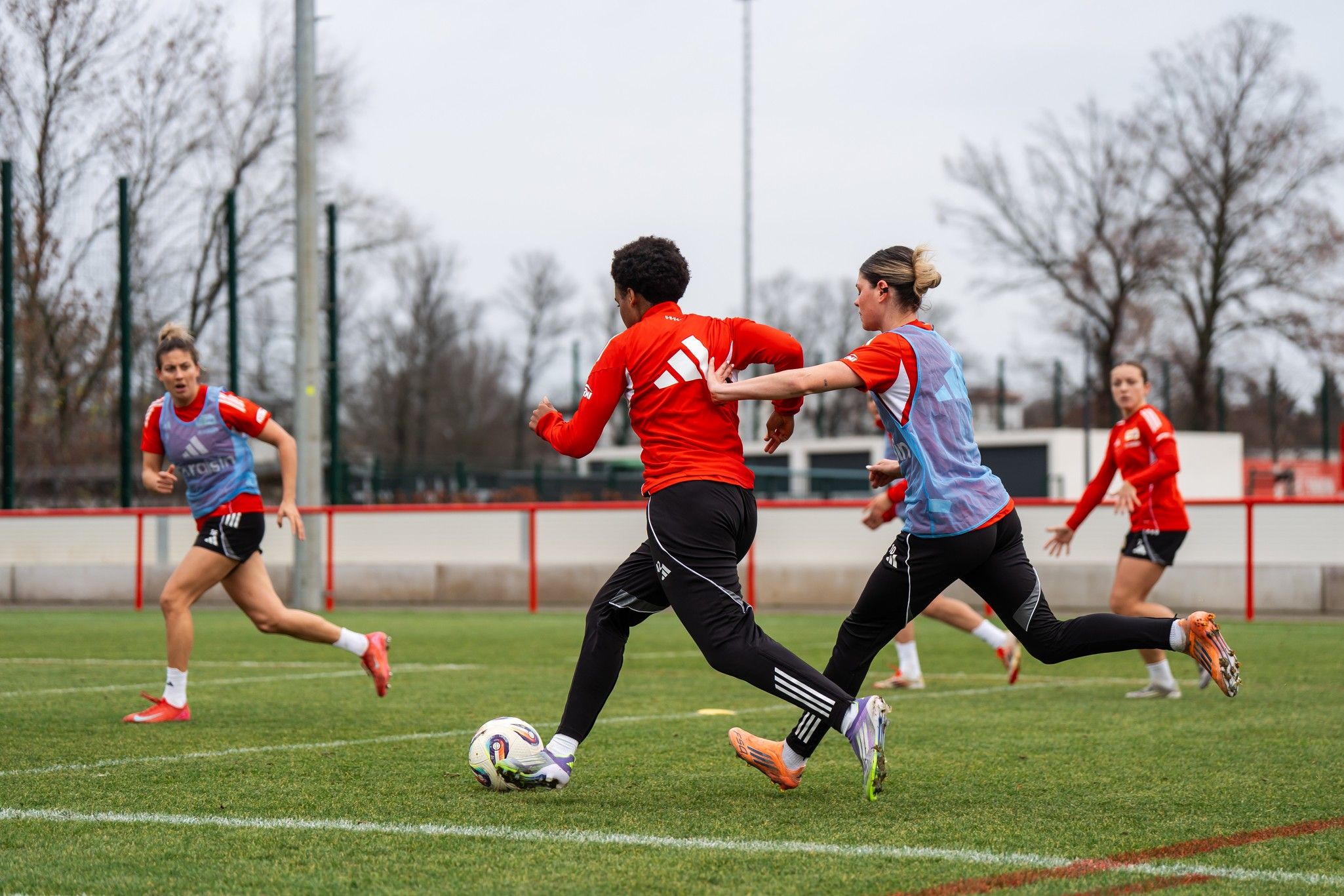 Zwei Fußballspielerinnen im roten Trikot trainieren auf einem grünen Platz, während eine weitere Spielerin im Hintergrund zuschaut.