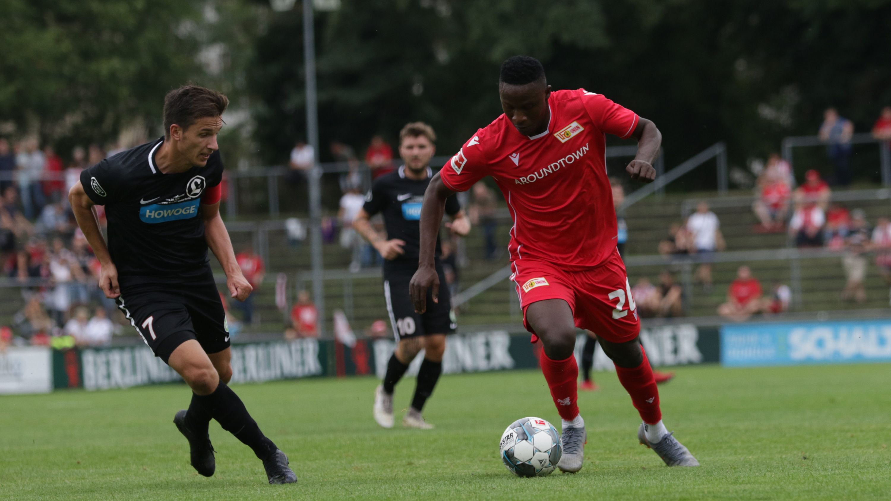 Zwei Fußballspieler auf dem Spielfeld, einer in rotem Trikot dribbelt, während der andere in schwarz ihn verfolgt.