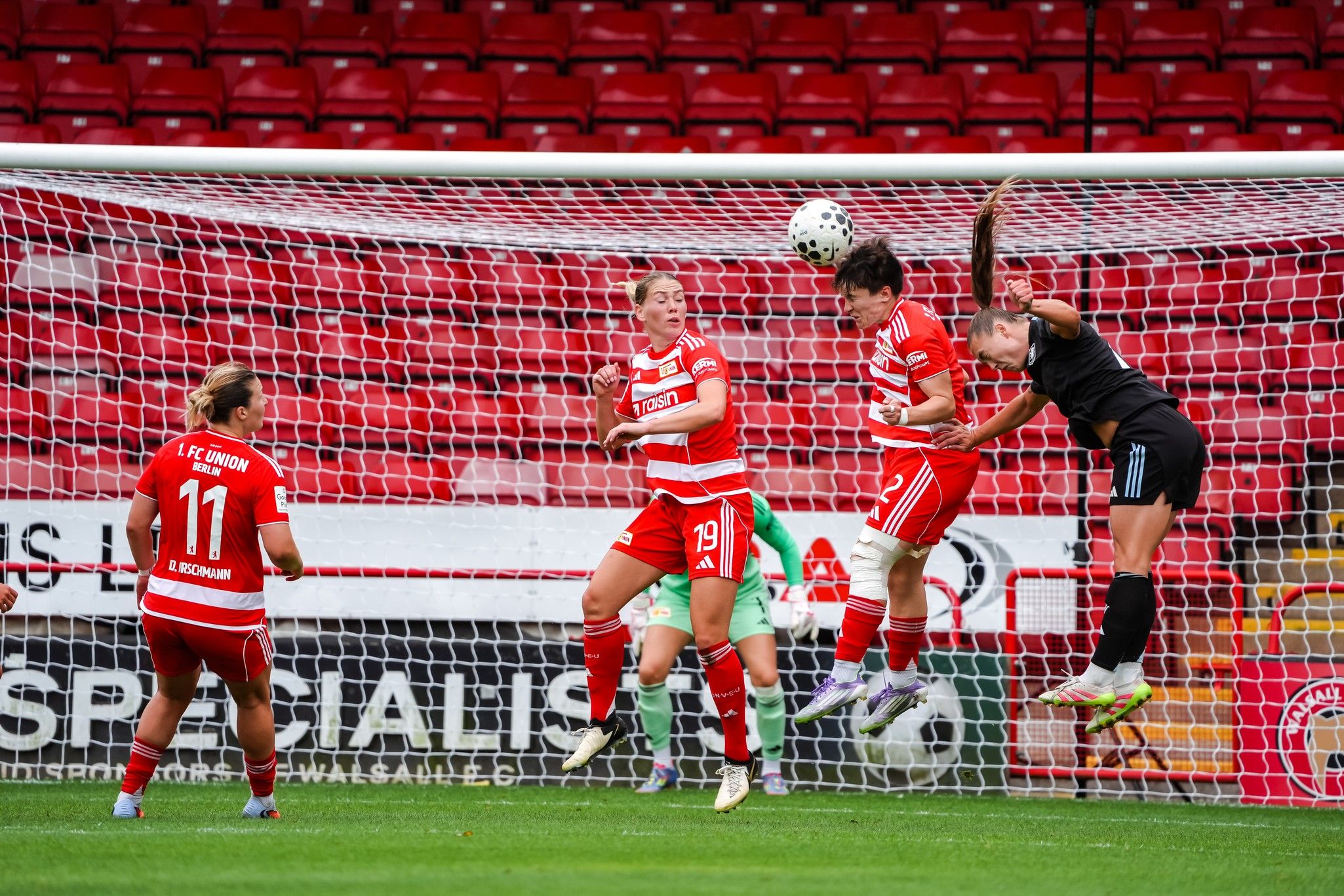 Spielszene im Fußball: Eine Spielerin springt hoch, um einen Ball zu köpfen, während ihre Mitspielerinnen und die Torhüterin zuschauen.