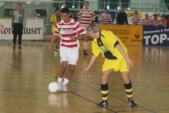 Zwei Fußballspieler in unterschiedlich farbigen Trikots spielen in einer Halle. Der Spieler in Rot und Weiß hat den Ball.