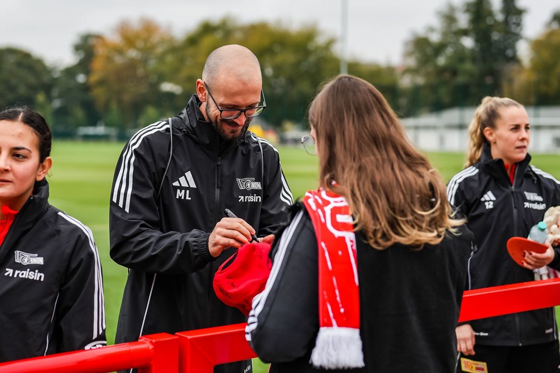 Personen in Sportbekleidung stehen an einem Zaun und interagieren, während im Hintergrund ein Fußballfeld zu sehen ist.