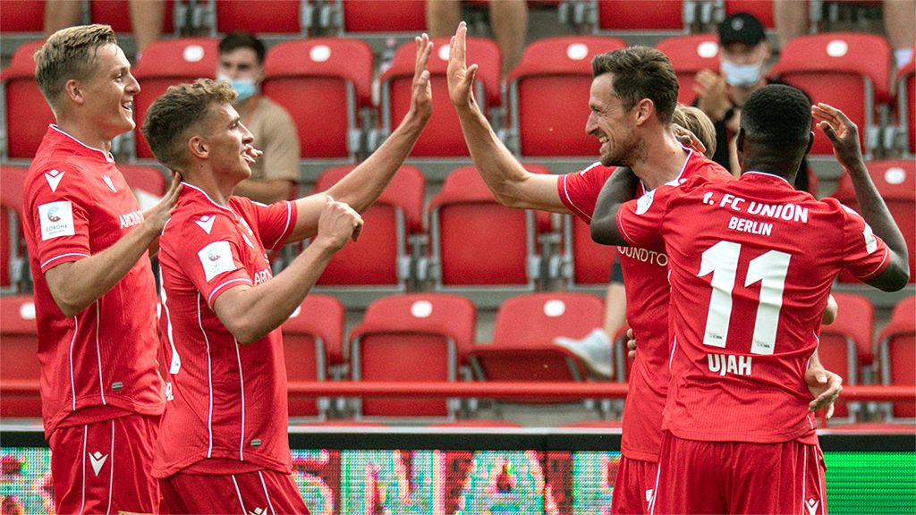 Five soccer players in red jerseys celebrate a goal together in a stadium.