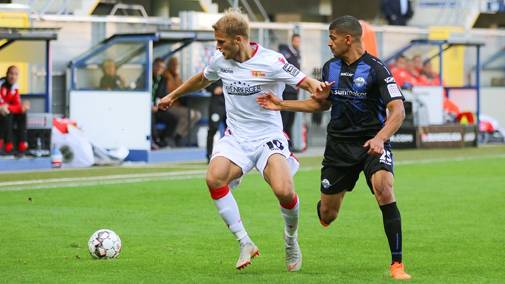 Ein Fußballspieler im weißen Trikot dribbelt den Ball, während ein Spieler im schwarzen Trikot ihn verteidigt.
