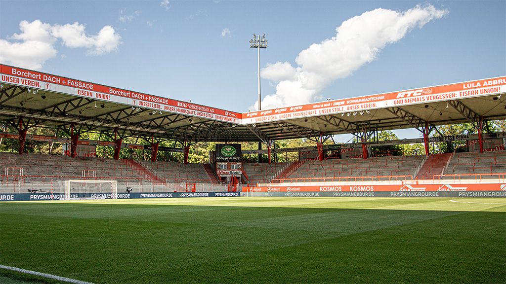 Leeres Fußballstadion mit Rasen, Tribünen und Werbebannern unter einem blauen Himmel.