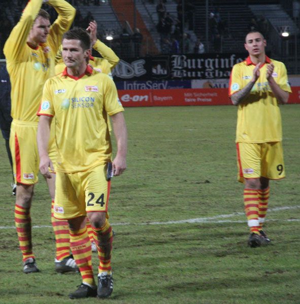 Drei Fußballspieler in gelben Trikots mit roten und gelben Streifen applaudieren auf dem Platz nach einem Spiel.