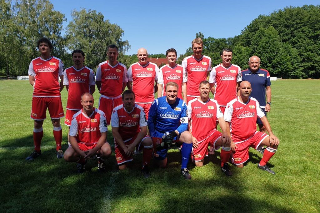 Elf Männer in roten Fußballtrikots stehen und hocken auf einem Sportfeld, umgeben von Bäumen an einem sonnigen Tag.