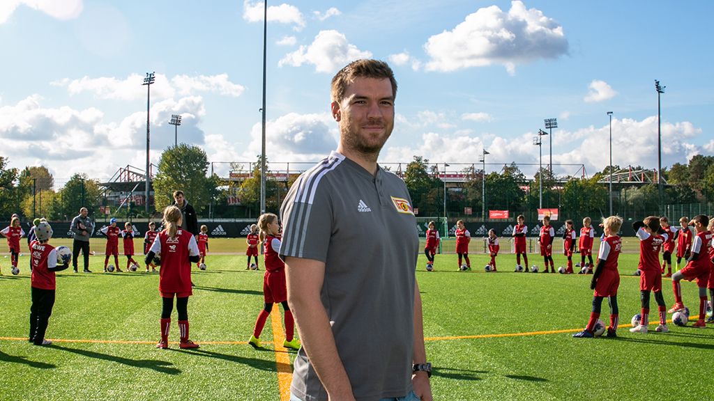 Trainer steht auf einem Fußballfeld, umgeben von jugendlichen Spielern in roten Trikots, die in einer Reihe aufgestellt sind.