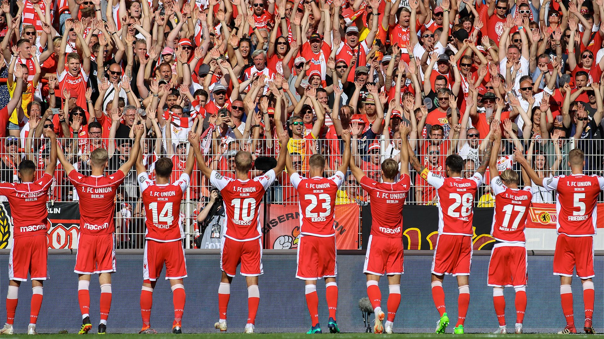 Players of a football team stand with their backs to the camera and enthusiastically wave to cheering fans in the stadium.