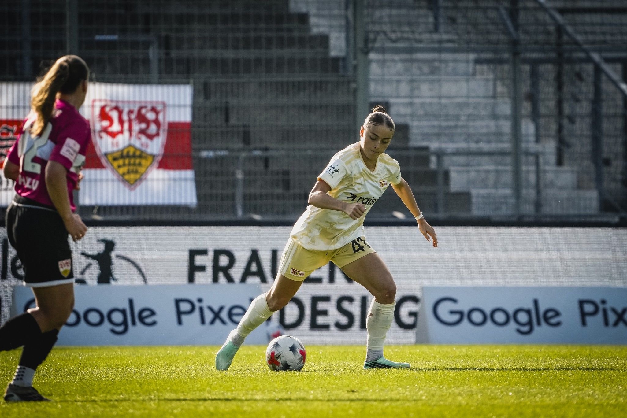 Eine Fußballspielerin in gelbem Trikot dribbelt den Ball auf einem Spielfeld vor einem Torwart und einem Vereinsbanner im Hintergrund.