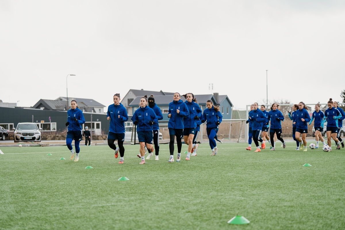 Eine Gruppe von Sportlern in blauen Trainingsanzügen joggt auf einem Rasenplatz, umgeben von Gebäuden und einer grauen Himmel.