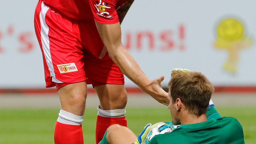 Ein Spieler in rotem Trikot hilft einem am Boden liegenden Torwart in grüner Ausrüstung auf.