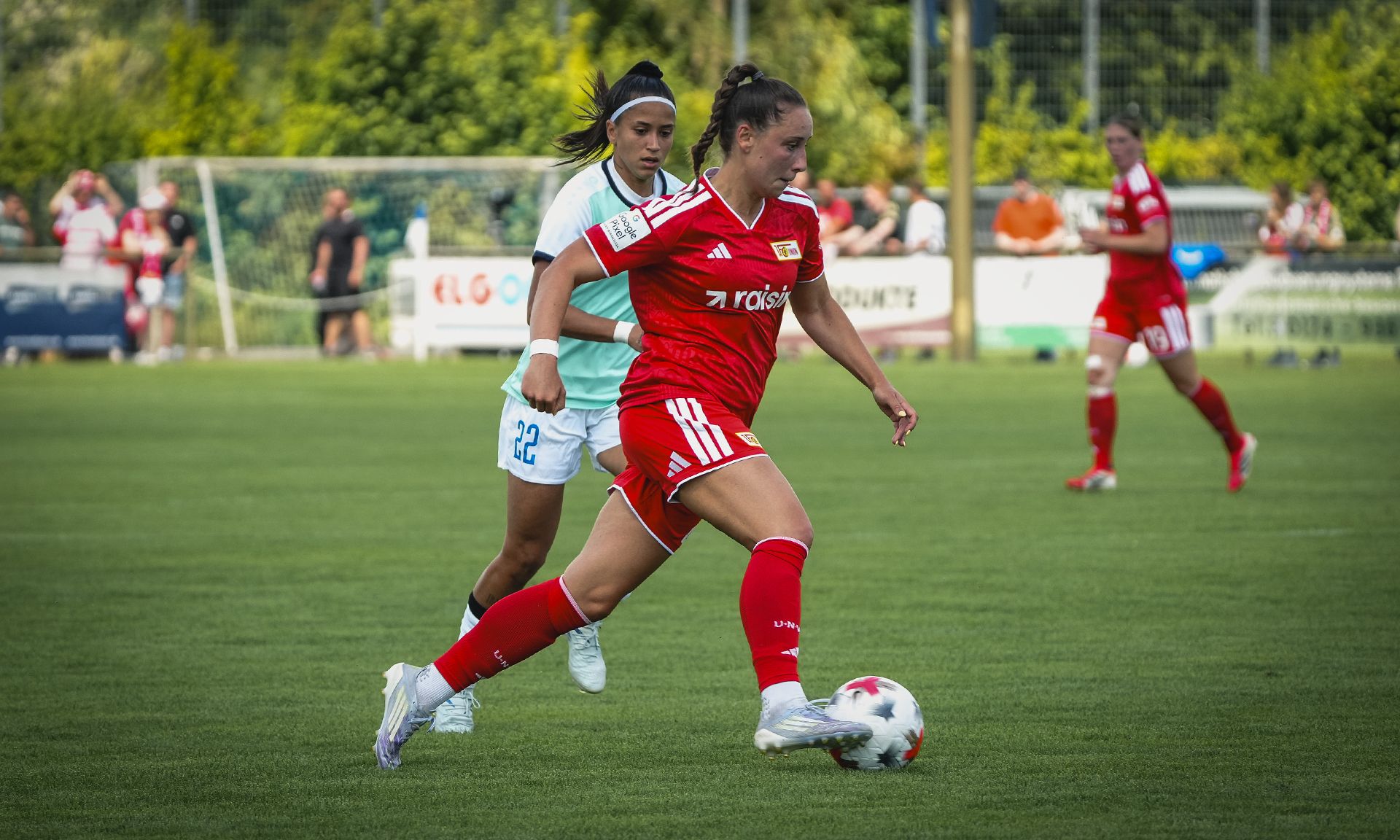 Eine Spielerin in rotem Trikot dribbelt den Ball, während eine Gegenspielerin in weißem Trikot verfolgt. Hintergrund mit Zuschauer und Fußballfeld.