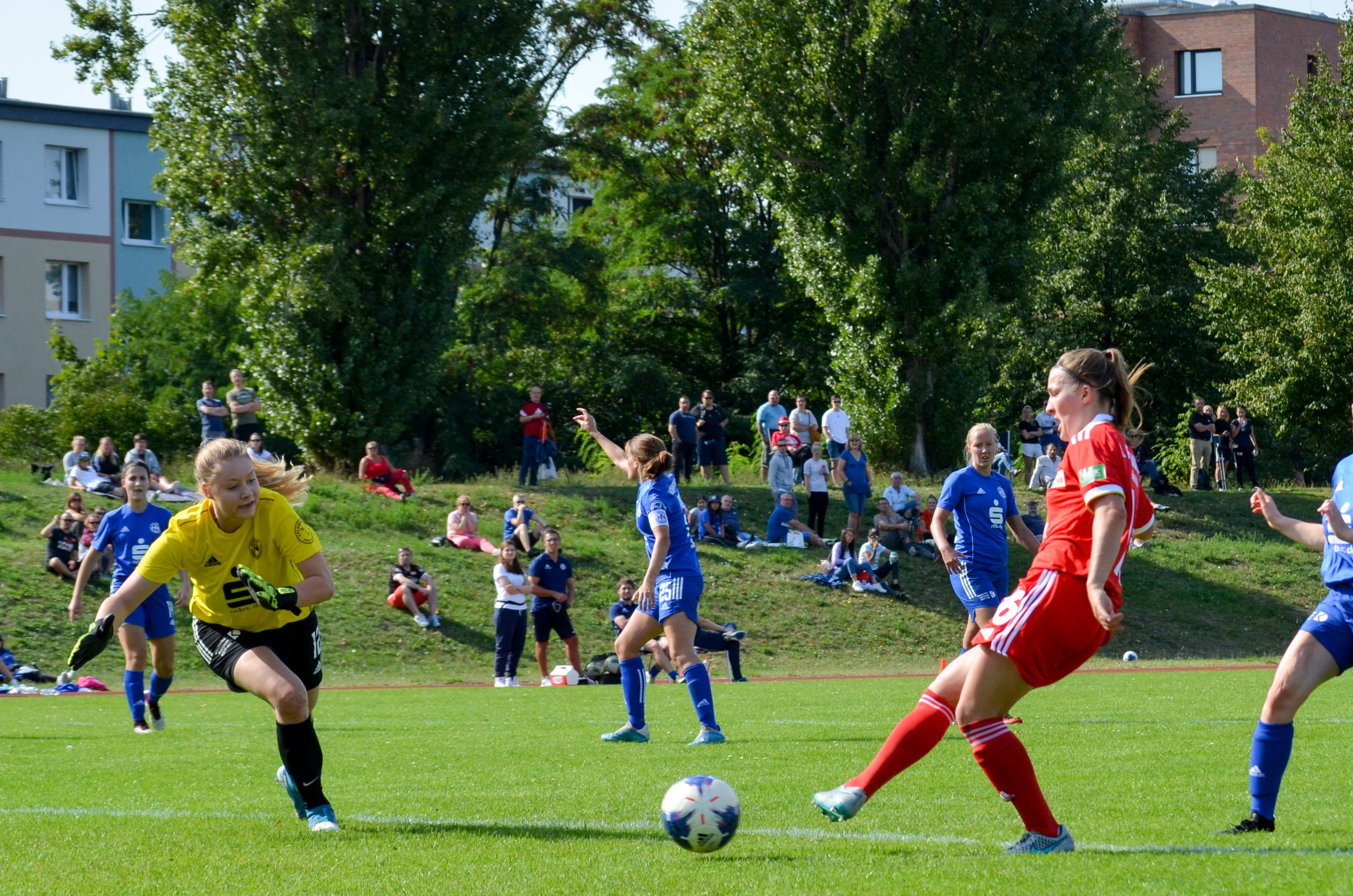 Eine Fußballspielszene mit einer Torhüterin in Gelb, die einen Ball abwehrt, während Spielerinnen in Blau und Rot auf dem Feld agieren.